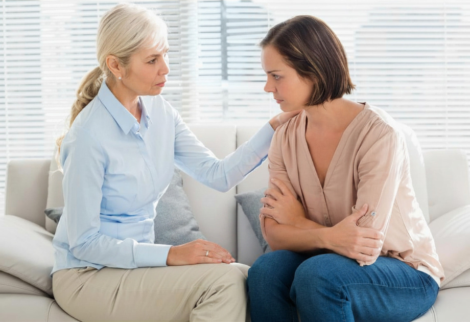 an older woman and younger woman sitting on a couch having a hard conversation. The older woman has her hand on the other woman's shoulder